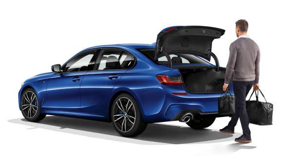 BMW 3. sērijas Sedan (G20), automātiskas bagāžas nodalījuma durvis. Trīs ceturtdaļu skats no aizmugures uz BMW 3. sērijas Sedan (G20) ar vadītāju, kurš ar savu kāju automātiski atver bagāžas nodalījuma durvis.