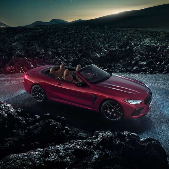 BMW M8 M Competition Cabrio, trīs ceturtdaļu skats no sāniem. BMW M8 Competition Cabrio metāliskā Motegi Red krāsā ar nolaistu mīkstā seguma jumtu, trīs ceturtdaļu skats no sāniem, stāvošs automobilis uz asfalta seguma naktī.