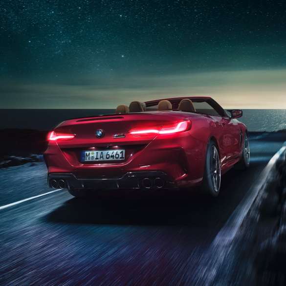 BMW M8 M Competition Cabrio, trīs ceturtdaļu skats no aizmugures. BMW M8 Competition Cabrio metāliskā Motegi Red krāsā ar nolaistu mīkstā seguma jumtu, trīs ceturtdaļu skats no aizmugures, braucošs automobilis pa piekrastes ceļu naktī.