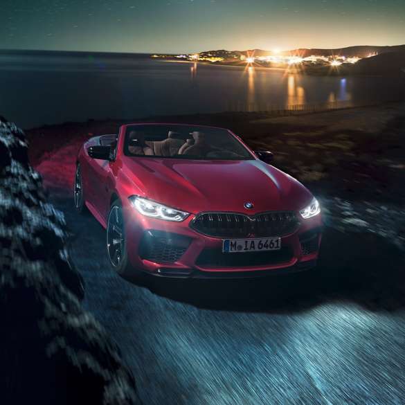 BMW M8 M Competition Cabrio, trīs ceturtdaļu skats no priekšpuses. BMW M8 Competition Cabrio metāliskā Motegi Red krāsā ar nolaistu mīkstā seguma jumtu, trīs ceturtdaļu skats no priekšpuses, braucošs automobilis pa piekrastes ceļu naktī.