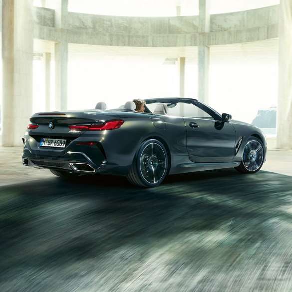 BMW M850i xDrive Cabrio, trīs ceturtdaļu skats no aizmugures. BMW M850i xDrive Competition Cabrio metāliskā Dravit Grey krāsā ar nolaistu mīkstā seguma jumtu, trīs ceturtdaļu skats no aizmugures uz automobili, kas brauc cauri betona ēkai.