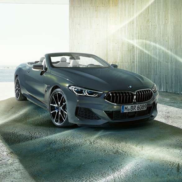 BMW M850i xDrive Cabrio, trīs ceturtdaļu skats no priekšpuses. BMW M850i xDrive Competition Cabrio metāliskā Dravit Grey krāsā ar nolaistu mīkstā seguma jumtu, trīs ceturtdaļu skats no sāniem, stāvošs automobilis uz piekrastes fona.