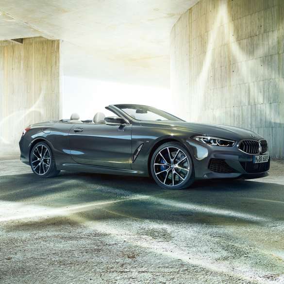 BMW M850i xDrive Cabrio, trīs ceturtdaļu skats no sāna. BMW M850i xDrive Competition Cabrio metāliskā Dravit Grey krāsā ar nolaistu mīkstā seguma jumtu, trīs ceturtdaļu skats no sāniem, stāvošs automobilis uz betona ēkas fona.
