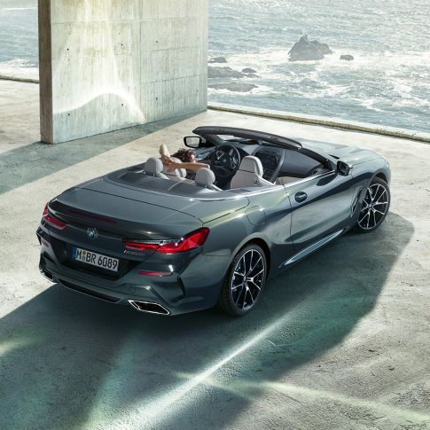 BMW M850i M Cabrio, skats no putna lidojuma. BMW M850i xDrive Cabrio metāliskā Dravit Grey krāsā ar nolaistu mīkstā seguma jumtu, skats no putna lidojuma, stāvēšanai novietots automobilis betona ēkā uz okeāna fona.