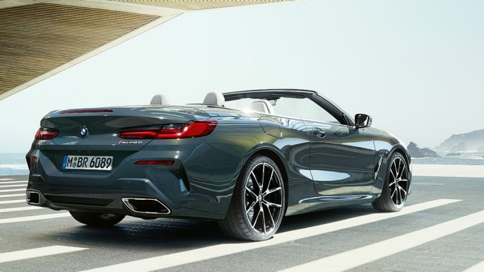 BMW M850i xDrive Cabrio, trīs ceturtdaļu skats no aizmugures. BMW M850i xDrive Competition Cabrio metāliskā Dravit Grey krāsā ar nolaistu mīkstā seguma jumtu, trīs ceturtdaļu skats no aizmugures uz novietotu automobili uz okeāna ainava fona.