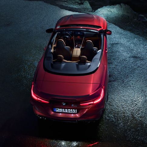 BMW M8 M Competition Cabrio, skats no putna lidojuma. BMW M8 Competition Cabrio metāliskā Motegi Red krāsā ar nolaistu mīkstā seguma jumtu, skats no putna lidojuma, stāvošs automobilis uz asfalta seguma naktī.