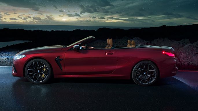 BMW M8 Competition Cabrio, skats no sāniem. BMW M8 Competition Cabrio metāliskā Motegi Red krāsā ar nolaistu mīkstā seguma jumtu, skats no sāniem, stāvošs automobilis ar zem zvaigžņotām debesīm.