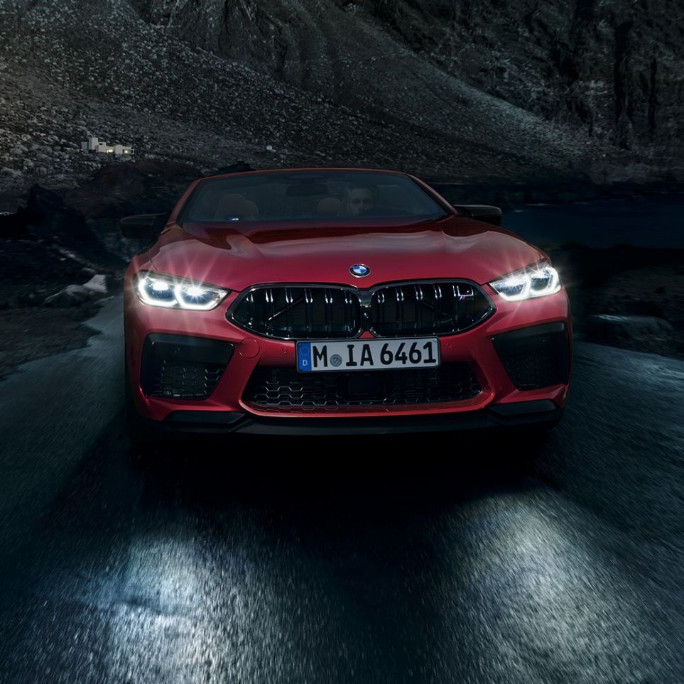BMW M8 Competition Cabrio, skats no priekšpuses. BMW M8 Competition Cabrio metāliskā Motegi Red krāsā ar ieslēgtiem priekšējiem lukturiem, skats no priekšpuses, braucošs automobilis naktī.