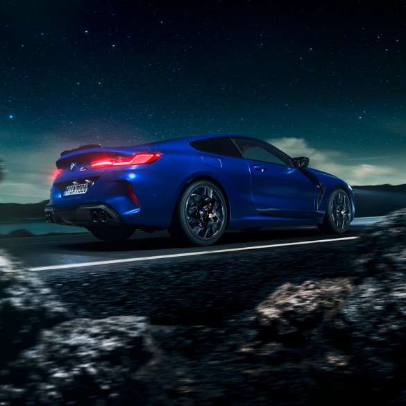 BMW M8 M Competition Coupé, trīs ceturtdaļu skats no sāniem. BMW M8 Competition Coupé metāliskā Marina Bay Blue krāsā, trīs ceturtdaļu skatā no priekšpuses uz braucošu automobili uz zvaigžņotas debess fona.