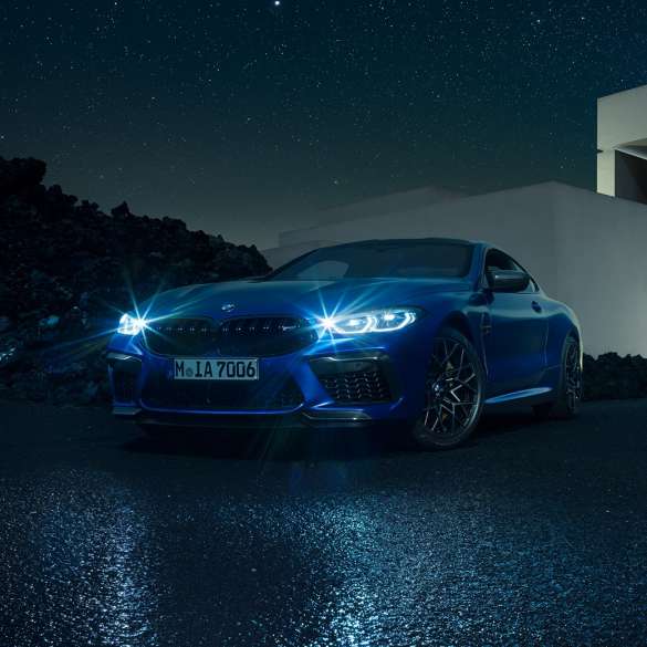 BMW M8 M Competition Coupé, trīs ceturtdaļu skats no priekšpuses. BMW M8 Competition Coupé metāliskā Marina Bay Blue krāsā ar ieslēgtiem LED priekšējiem lukturiem, trīs ceturtdaļu skatā no priekšpuses uz villas priekšā novietotu automobili naktī.