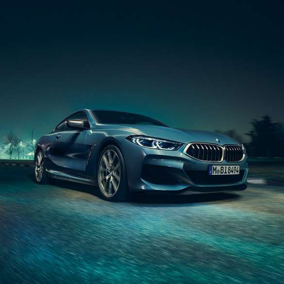 BMW M850i xDrive Coupé, priekšpuse trīs ceturtdaļu skatā no kreisās puses. BMW M850i xDrive Coupé Coupé metāliskā Barcelona Blue krāsā, priekšpuse trīs ceturtdaļu skatā no kreisās puses ar braucošu automobili naktī.