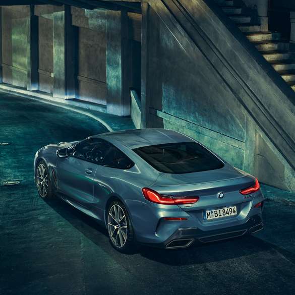 BMW M850i xDrive Coupé, aizmugure trīs ceturtdaļu skatā no kreisās puses. BMW M850i xDrive Coupé Coupé metāliskā Barcelona Blue krāsā, priekšpuse trīs ceturtdaļu skatā no kreisās puses ar stāvošu automobili naktī.