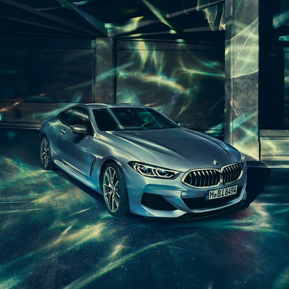 BMW M850i xDrive Coupé, priekšpuse trīs ceturtdaļu skatā no kreisās puses. BMW M850i xDrive metāliskā Barcelona Blue krāsā, trīs ceturtdaļu skats uz stāvēšanai novietotu automobili no priekšpuses uz ēkas fona.