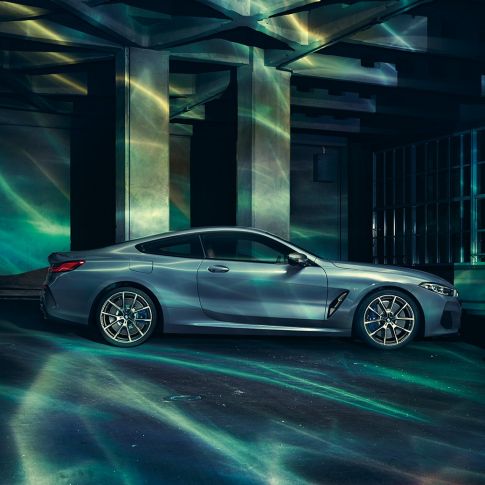 BMW M850i xDrive Coupé, skats no sāniem. BMW M850i xDrive Coupé metāliskā Barcelona Blue krāsā, skats no sāniem uz novietotu automobili uz ēkas fona.