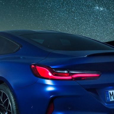 M aizmugurējais spoileris BMW M8 Competition Coupé metāliskā Marina Bay Blue krāsā, naksnīgs plecu daļas tuvplāns ar M aizmugurējo oglekļa spoileri.