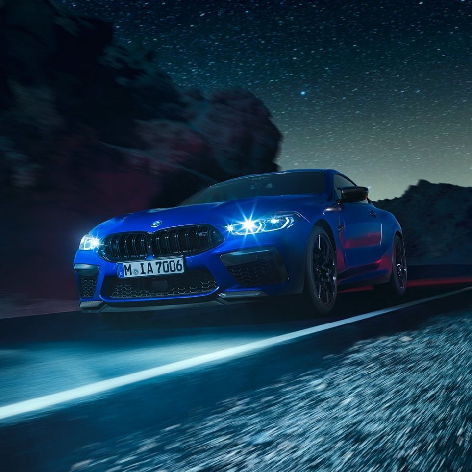 Braucošs BMW M8 M Competition Coupé, trīs ceturtdaļu skats no priekšpuses. BMW M8 Competition Coupé metāliskā Marina Bay Blue krāsā ar iedegtiem LED priekšējiem lukturiem, trīs ceturtdaļu skatā no priekšpuses uz braucošu automobili naktī.