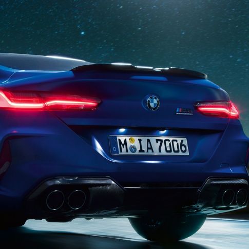 M difuzors Carbon Black krāsā. BMW M8 Competition Coupé metāliskā Marina Bay Blue krāsā, aizmugures tuvplāns naktī ar M difuzoru Carbon Black krāsā.
