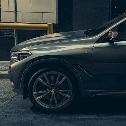 BMW X6 M50i skatā no sāna Vīrietis, kas atbalstās pret BMW X6 M50i skatā no sāna uz pilsētas fona.