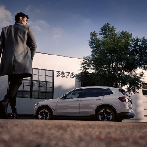 BMW iX3, G08, 2020, sānskats uz ēkas fona ar vīrieti BMW iX3, G08, 2020, metāliska Mineral White krāsa, sānskats uz ēkas fona ar vīrieti