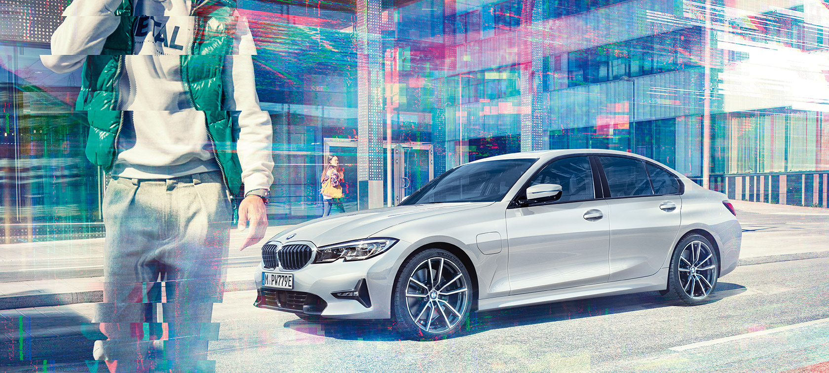 BMW 3. sērijas Sedan, skats no sāna uz BMW 330e. Trīs ceturtdaļu skats no sāniem uz stāvošu BMW 330e Sedan uz stiklotas ēkas un ejošu pasažieru fona.