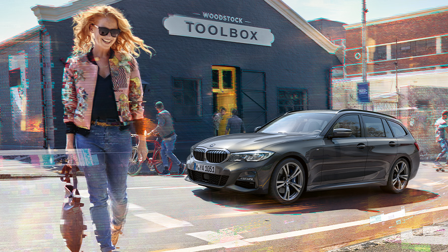 BMW 3. sērijas Touring (G21), Dravit Grey krāsa, trīs ceturtdaļu skats no priekšpuses uz sievieti ar kokapstrādes instrumentu veikalu fonā. Sieviete šķērso ielu uz BMW 3. sērijas Touring (G21) fona, kas atrodas pie kokapstrādes instrumentu veikala.