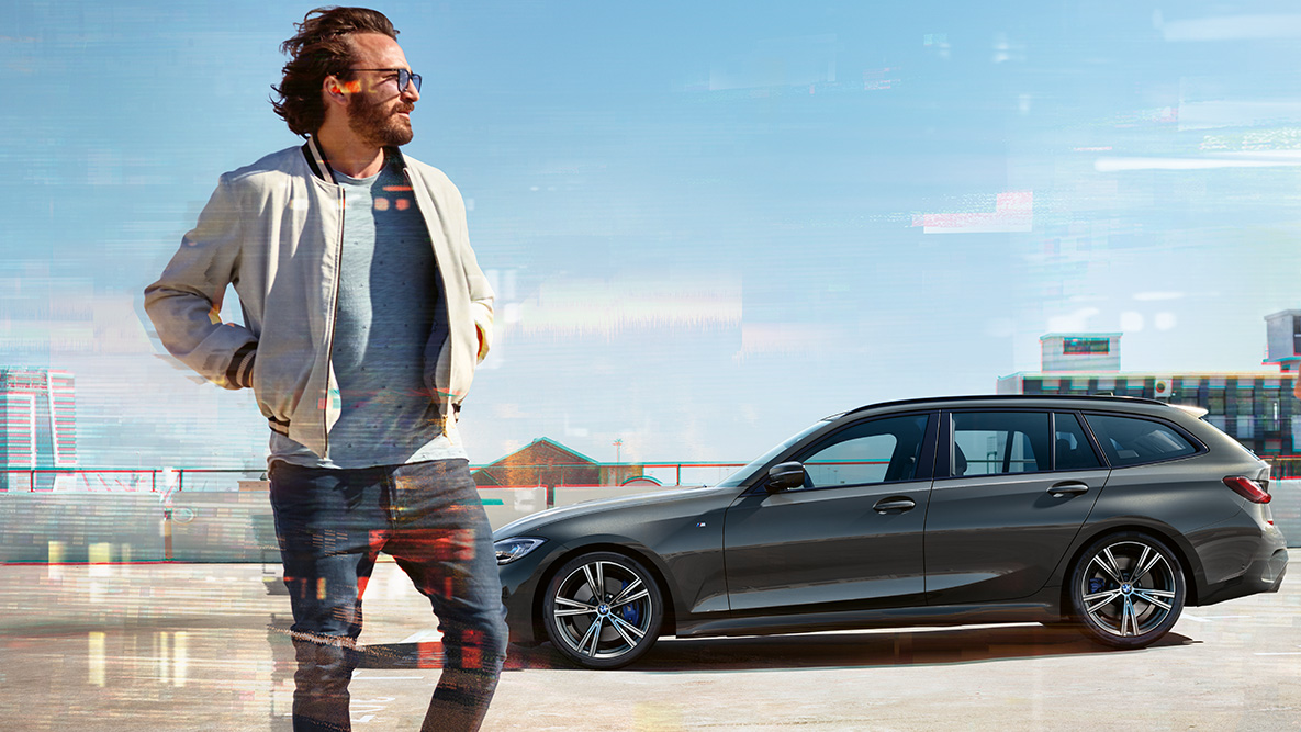 BMW 3. sērijas Touring (G21), trīs ceturtdaļu skats no priekšpuses dizaina novērtēšanai BMW 3. sērijas Touring (G21), Dravit Grey krāsa, trīs ceturtdaļu skats no priekšpuses uz braucošu automobili, šķērsojot tiltu, ar lauku apvidu fonā