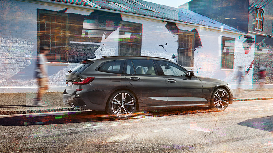 BMW 3. sērijas Touring (G21), Dravit Grey krāsa, septiņu astotdaļu aizmugures skats BMW 3. sērijas Touring (G21), Dravit Grey krāsa, septiņu astotdaļu skats no aizmugures uz braucošu automobili uz ēkas fona