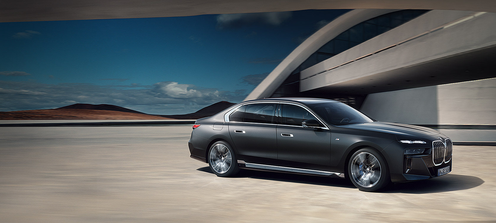 BMW 7. sērijas Sedan, G70, ICE, M sporta pakotne, metāliska Frozen Deep Grey krāsa, trīs ceturtdaļu priekšpuses skats, stāvošs automobilis uz ēkas fona