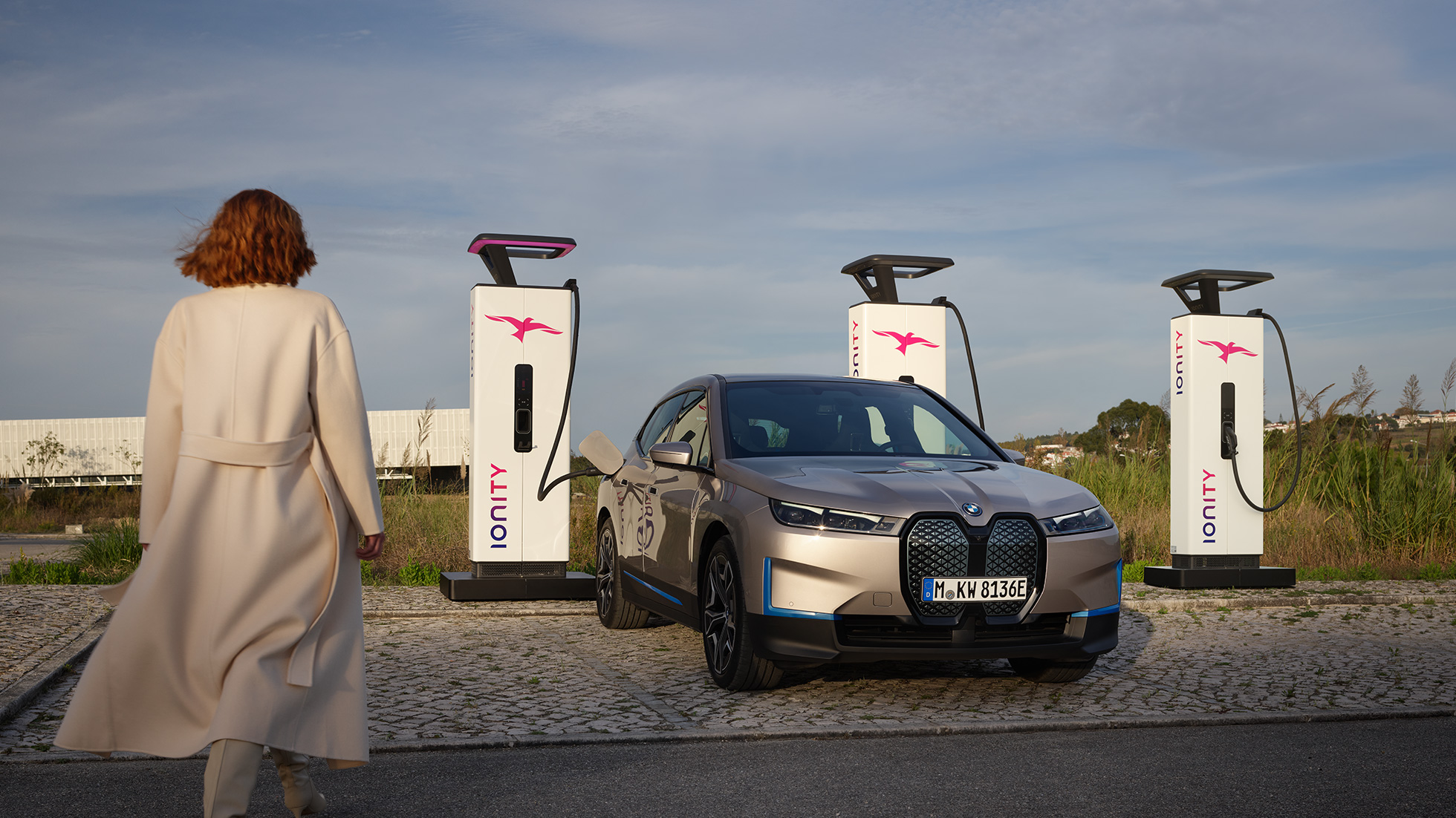 BMW iX, trīs ceturtdaļu priekšpuses skats uzlādes stacijā BMW iX, Cashmere Silver krāsa, i20, trīs ceturtdaļu priekšpuses skats uzlādes stacijā