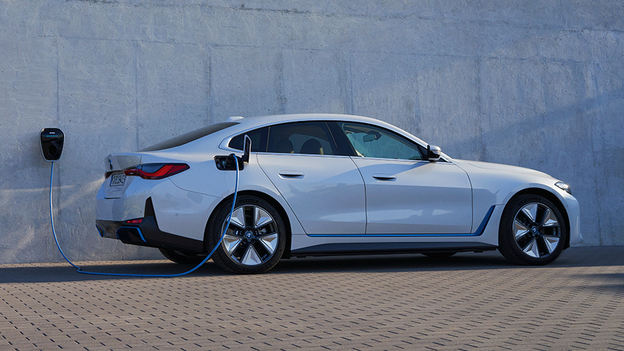 Sānskats ar BMW Wallbox BMW i4 eDrive40, G26, 2021 BMW i4 eDrive40, G26, 2021, Mineral White krāsa, sānskats uz ēkas sienas fona ar BMW Wallbox uzlādes staciju