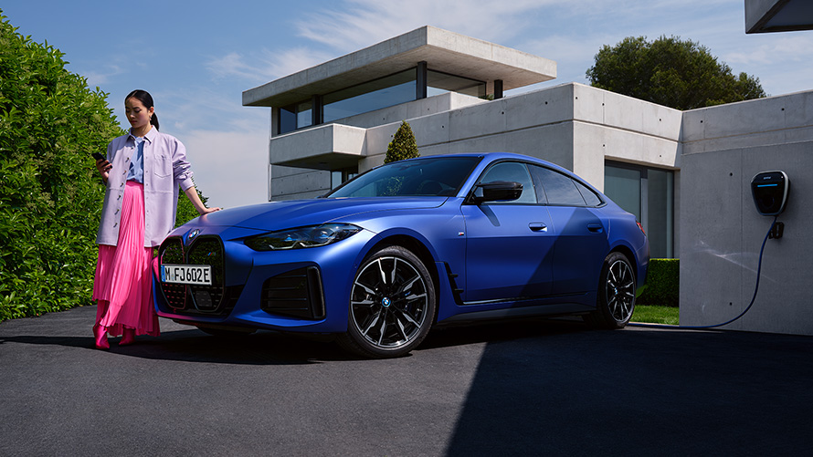 BMW i4 M50, G26, 2021, BMW Individual metāliska Frozen Portimao Blue krāsa, trīs ceturtdaļu priek&scaron;puses skats, stāvošs automobilis ar sievieti uz ēkas fona