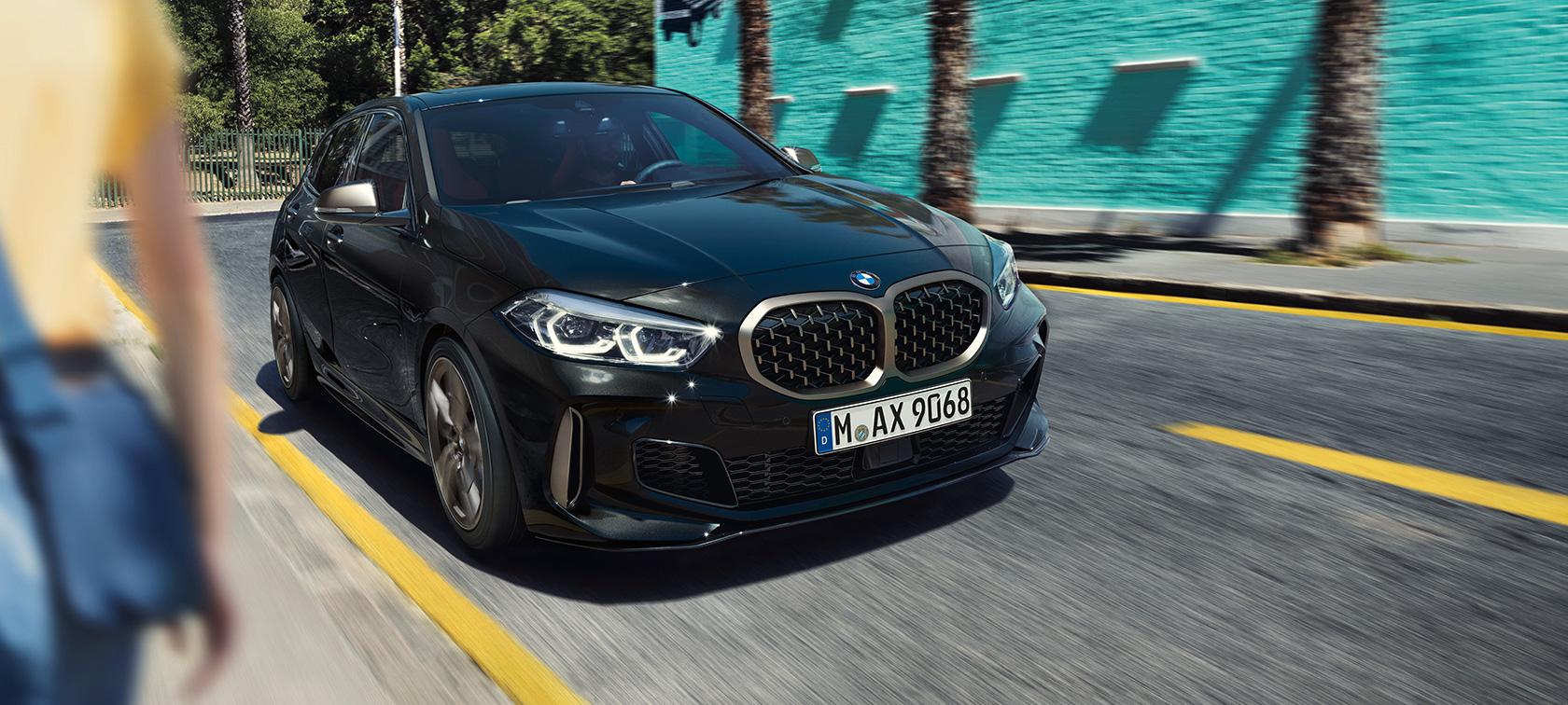 BMW M135i xDrive, trīs ceturtdaļu skats no priekšpuses. BMW M135i xDrive metāliskā Black Sapphire krāsā, trīs ceturtdaļu skats uz braucošu automobili no priekšpuses.