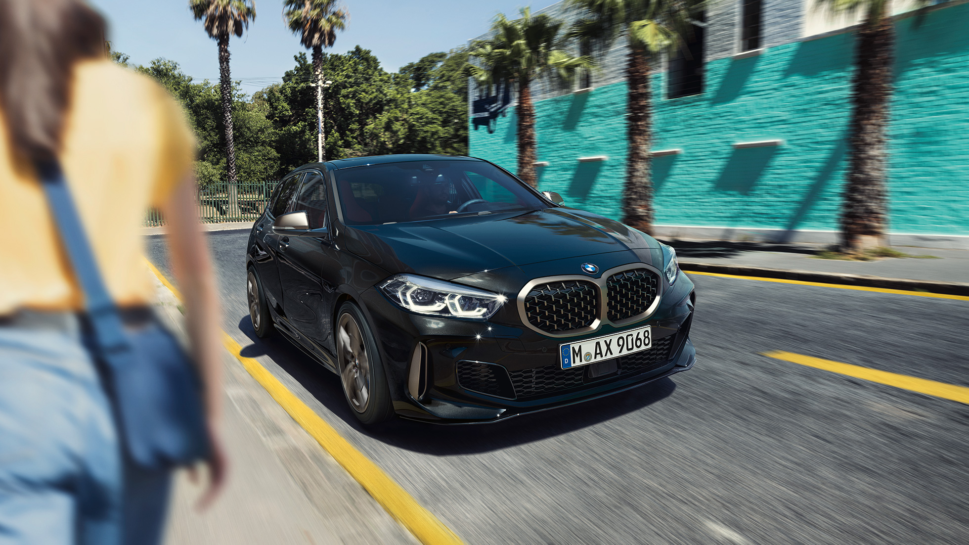 BMW M135i xDrive, trīs ceturtdaļu skats no priekšpuses. BMW M135i xDrive metāliskā Black Sapphire krāsā, trīs ceturtdaļu skatā uz braucošu automobili no priekšpuses ar sievieti priekšplānā.