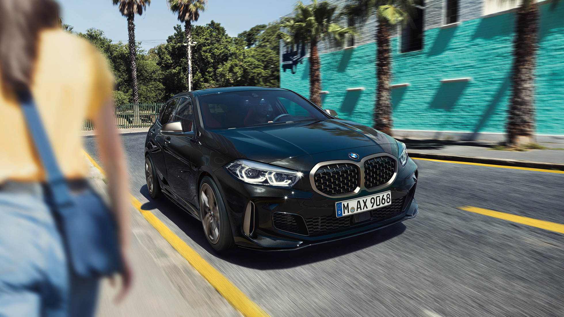 BMW M135i xDrive, trīs ceturtdaļu skats no priekšpuses. BMW M135i xDrive metāliskā Black Sapphire krāsā, trīs ceturtdaļu skatā uz braucošu automobili no priekšpuses ar sievieti priekšplānā.