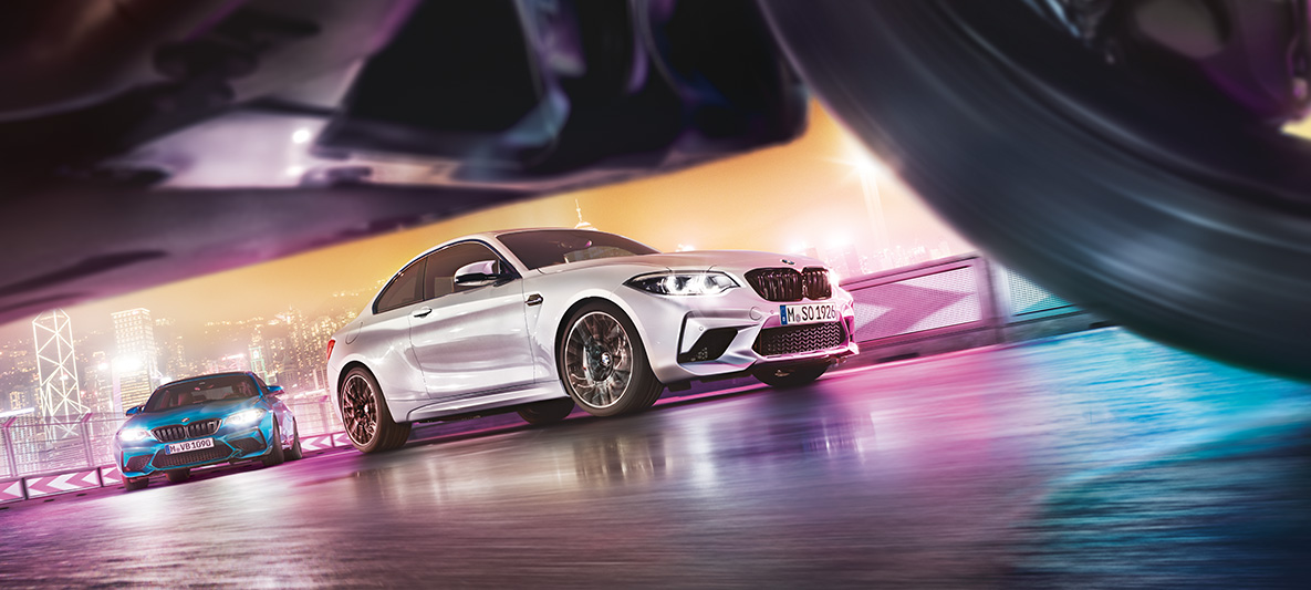 BMW M2 Competition uz atstarojoša asfalta. Divi BMW M2 Competition automobiļi sacīkšu trasē ar naksnīgu pilsētu fonā – ielas līmeņa skats.