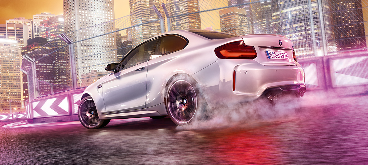 BMW M2 Competition uzņem ātrumu Balta BMW M2 Competition automobiļa drifta manevri sacīkšu trasē ar naksnīgu apvārsni fonā.