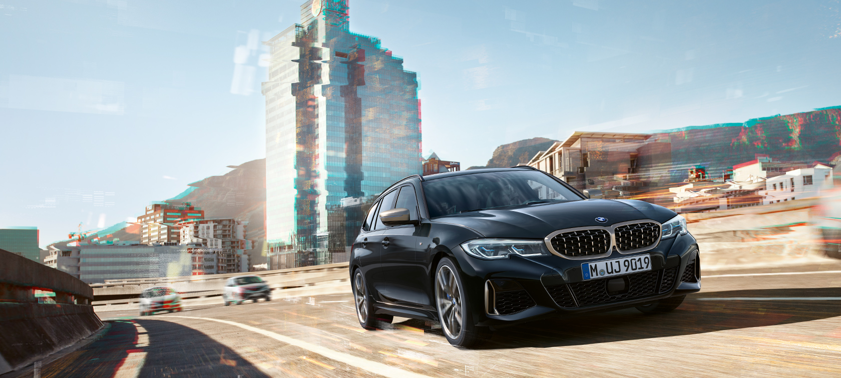 Trīs ceturtdaļu skats braucienā uz pilsētas fona BMW M340i xDrive Touring, trīs ceturtdaļu skats braucienā uz pilsētas fona