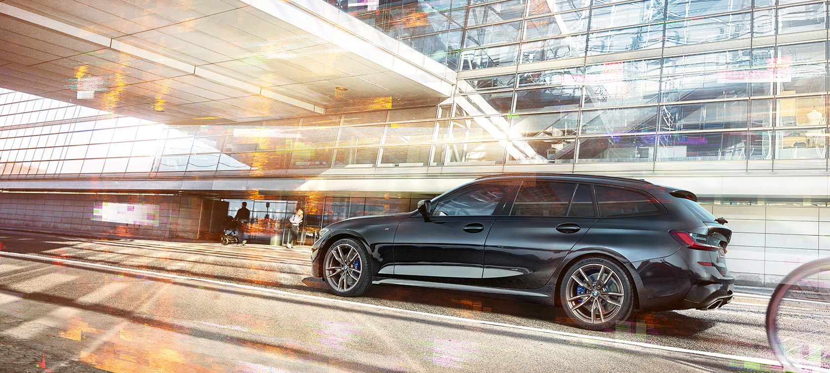 BMW M340i xDrive Touring, skats no sāna braucienā BMW M340i xDrive Touring, skats no sāna braucienā uz pilsētnieciskas stiklotas ēkas fona