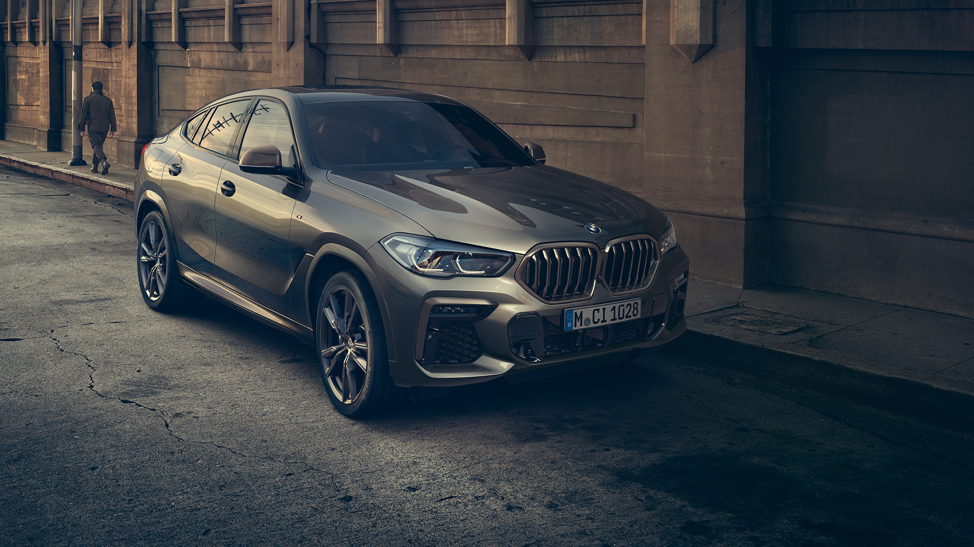 Novietots BMW X6 M50i. BMW X6 M50i trīs ceturtdaļu skats uz novietotu automobili no priekšpuses uz pilsētas fona.