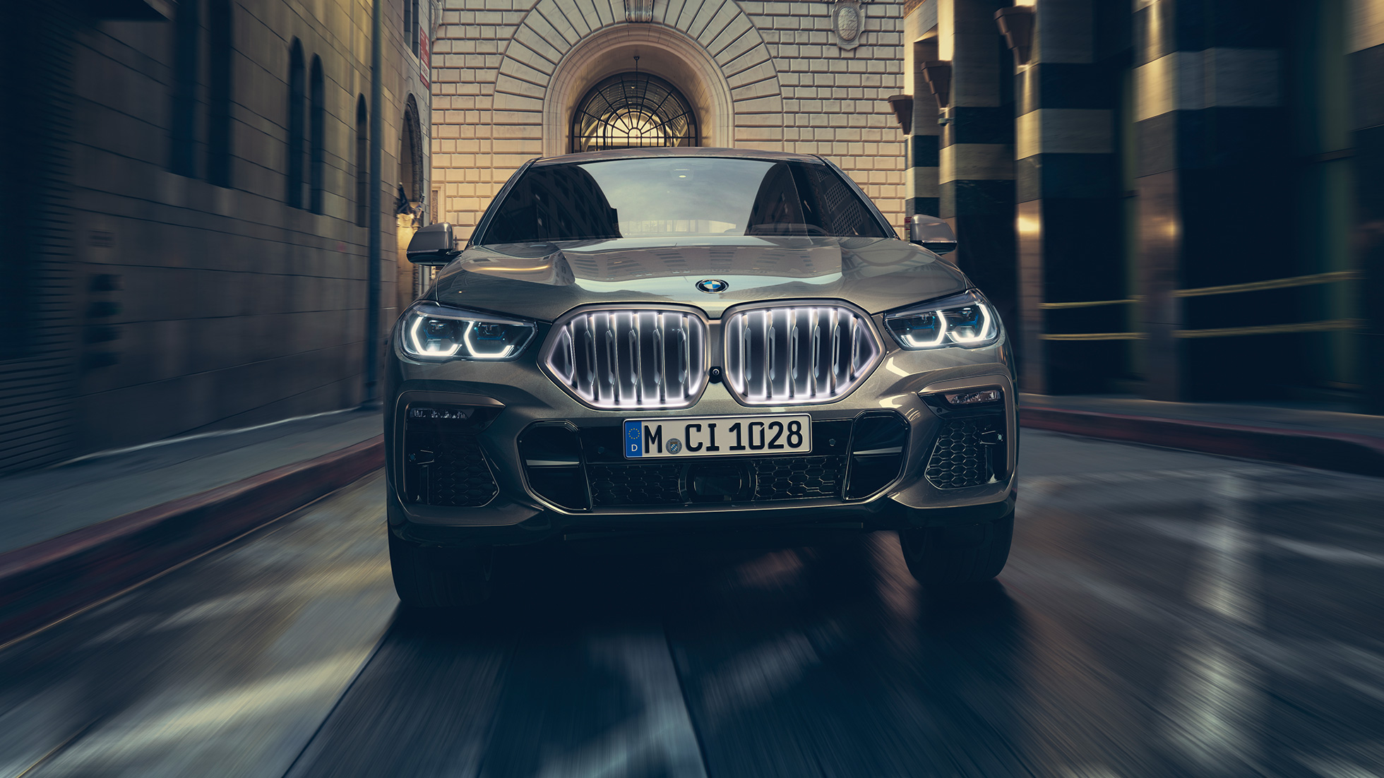 BMW X6 M50i ar BMW elipsveida režģi‚ “Iconic Glow”. BMW X6 M50i skats no aizmugures uz pilsētas fona.