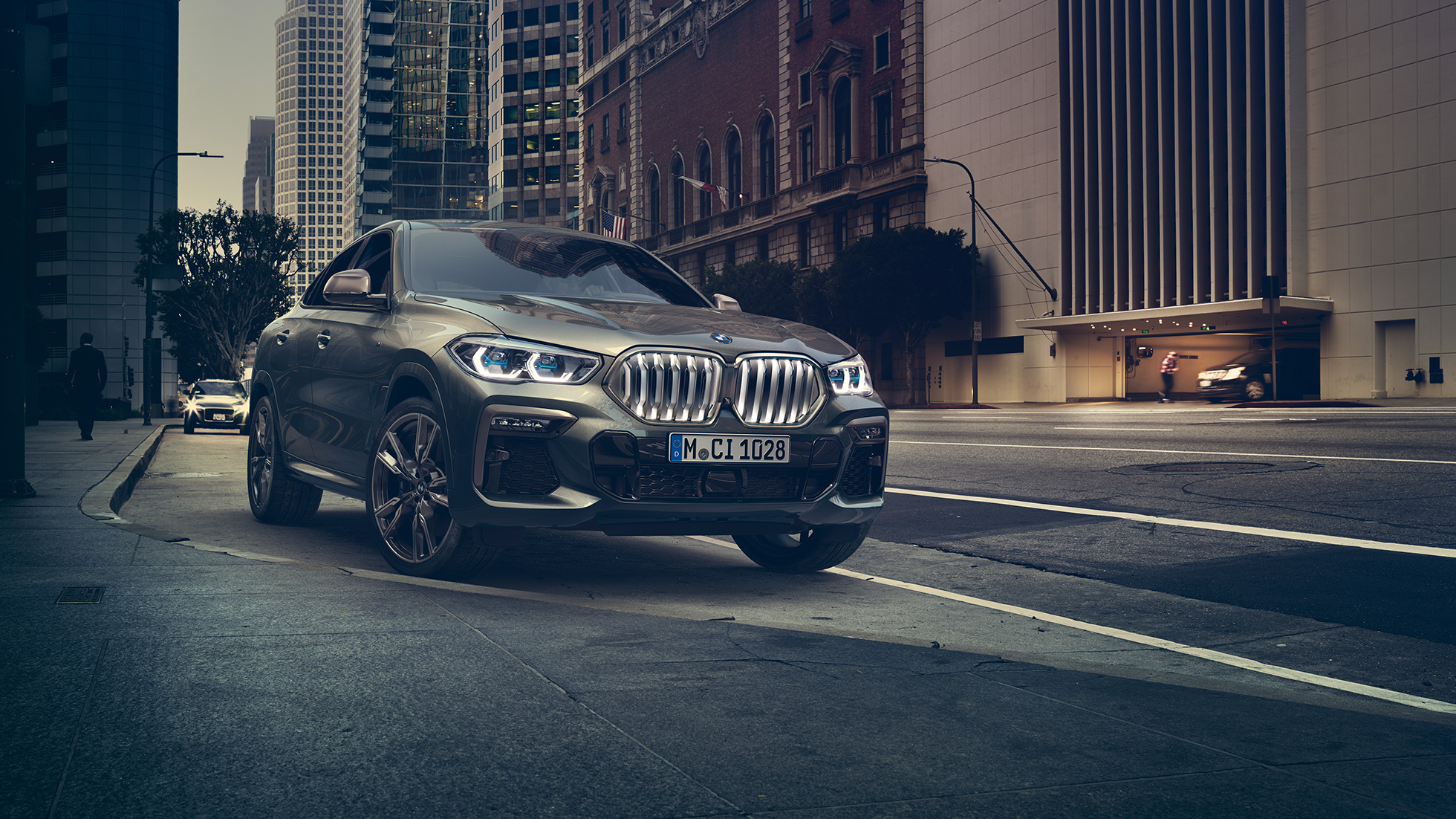 Novietots BMW X6 M50i. BMW X6 M50i trīs ceturtdaļu skats uz novietotu automobili no priekšpuses uz pilsētas fona.