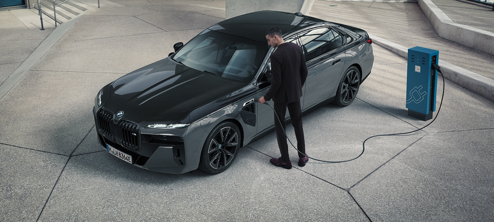 BMW M760e xDrive Sedan, G70, M Performance pakotne BMW M760e xDrive Sedan, G70, M Performance pakotne, BMW Individual divtoņu metālisks Black Sapphire krāsojums, BMW Individual metāliska Dravite Grey krāsa, trīs ceturtdaļu priekšpuses skats, vīrietis ar uzlādes kabeli blakus uzlādes stacijai