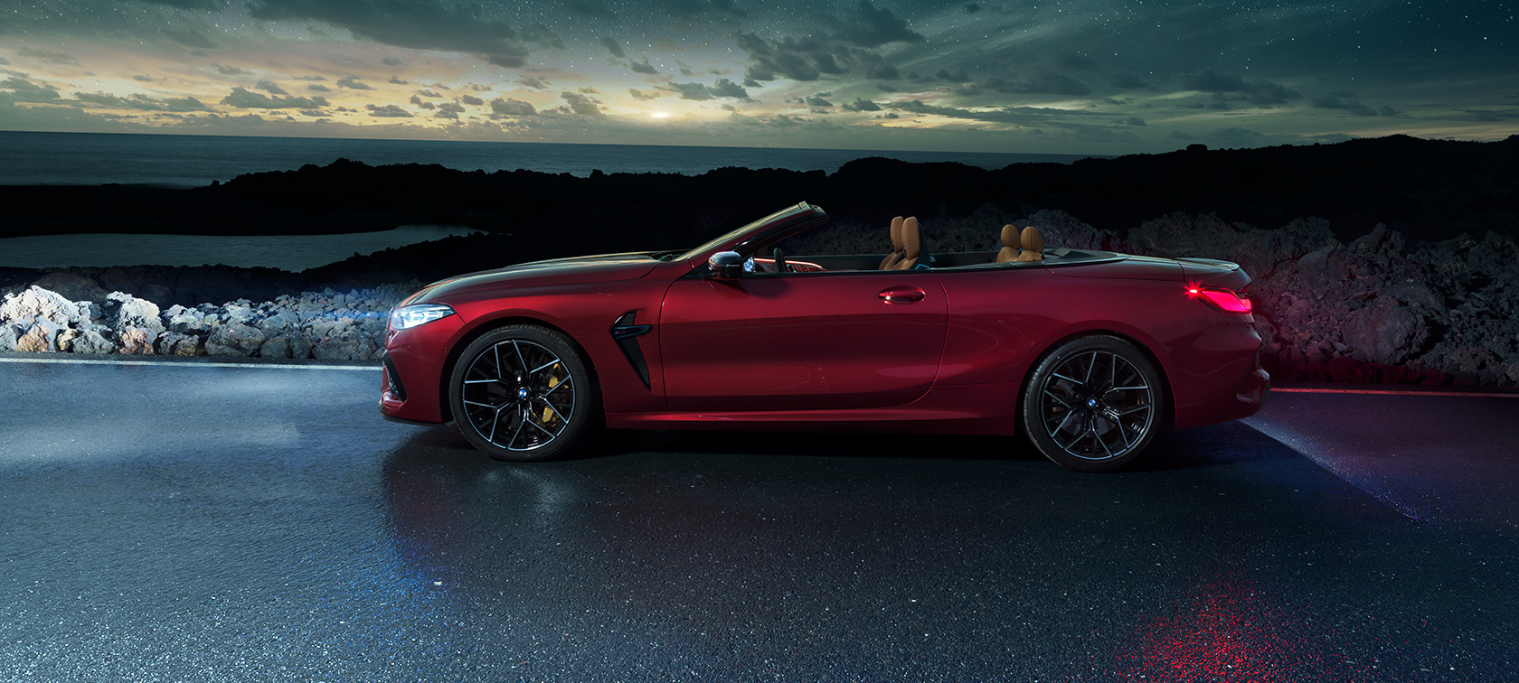BMW M8 Competition Cabrio, skats no sāniem. BMW M8 Competition Cabrio metāliskā Motegi Red krāsā ar nolaistu mīkstā seguma jumtu, skats no sāniem, piekrastē stāvošs automobilis ar zem zvaigžņotām debesīm.