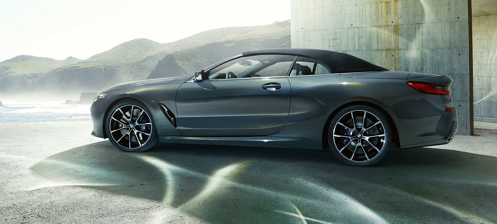 BMW M850i xDrive Cabrio, skats no sāniem. BMW M850i xDrive Cabrio metāliskā Dravit Grey krāsā ar nolaistu mīkstā seguma jumtu, skats no sāniem uz kalnu fona.