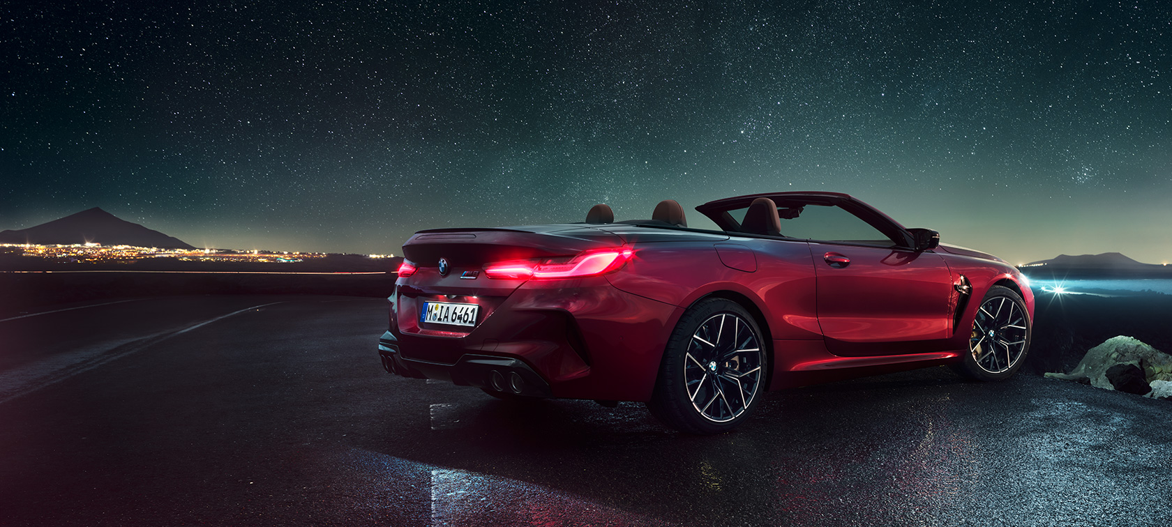 BMW M8 M Competition Cabrio, trīs ceturtdaļu skats no aizmugures. BMW M8 Competition Cabrio metāliskā Motegi Red krāsā ar nolaistu mīkstā seguma jumtu, aizmugures trīs ceturtdaļu skats no sāniem, stāvošs automobilis zem zvaigžņotas debess.
