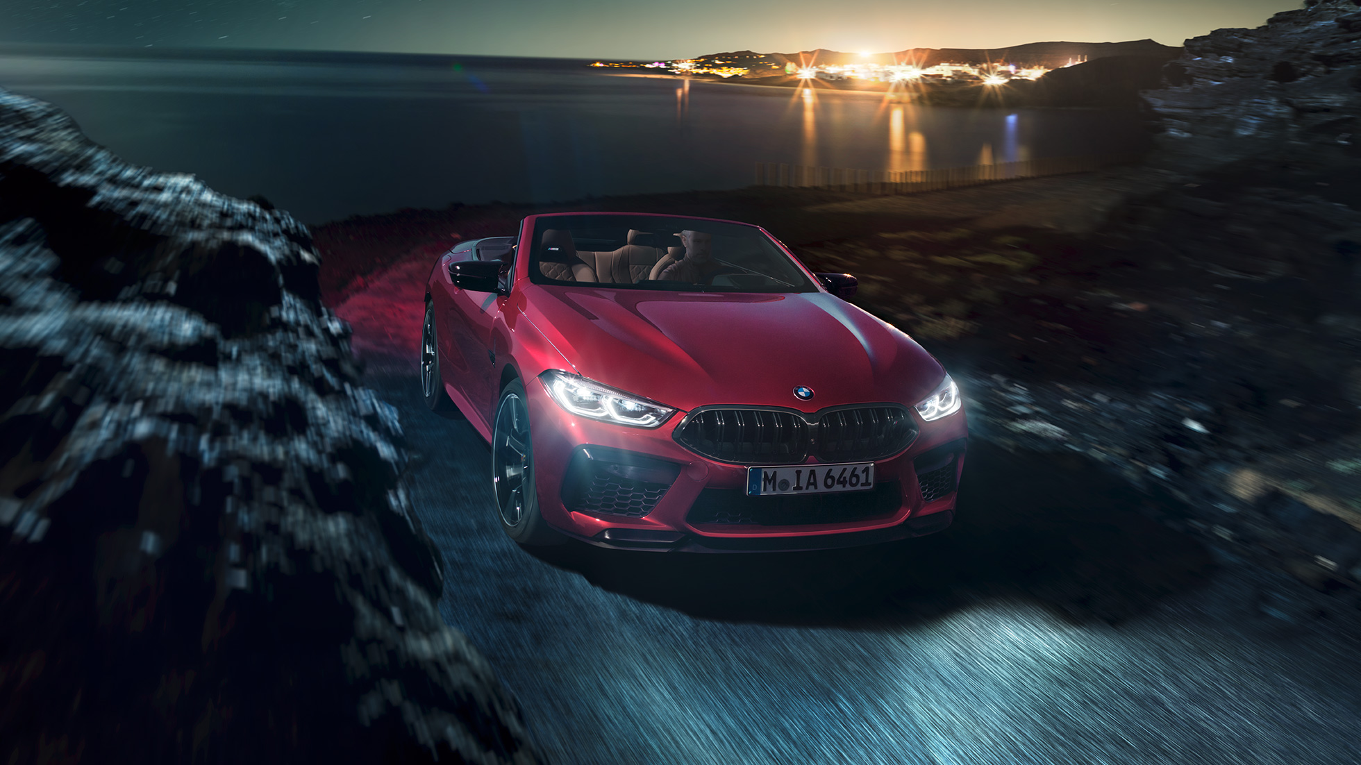 BMW M8 M Competition Cabrio, trīs ceturtdaļu skats no priekšpuses. BMW M8 Competition Cabrio metāliskā Motegi Red krāsā ar nolaistu mīkstā seguma jumtu, trīs ceturtdaļu skats no priekšpuses, braucošs automobilis pa piekrastes ceļu naktī.