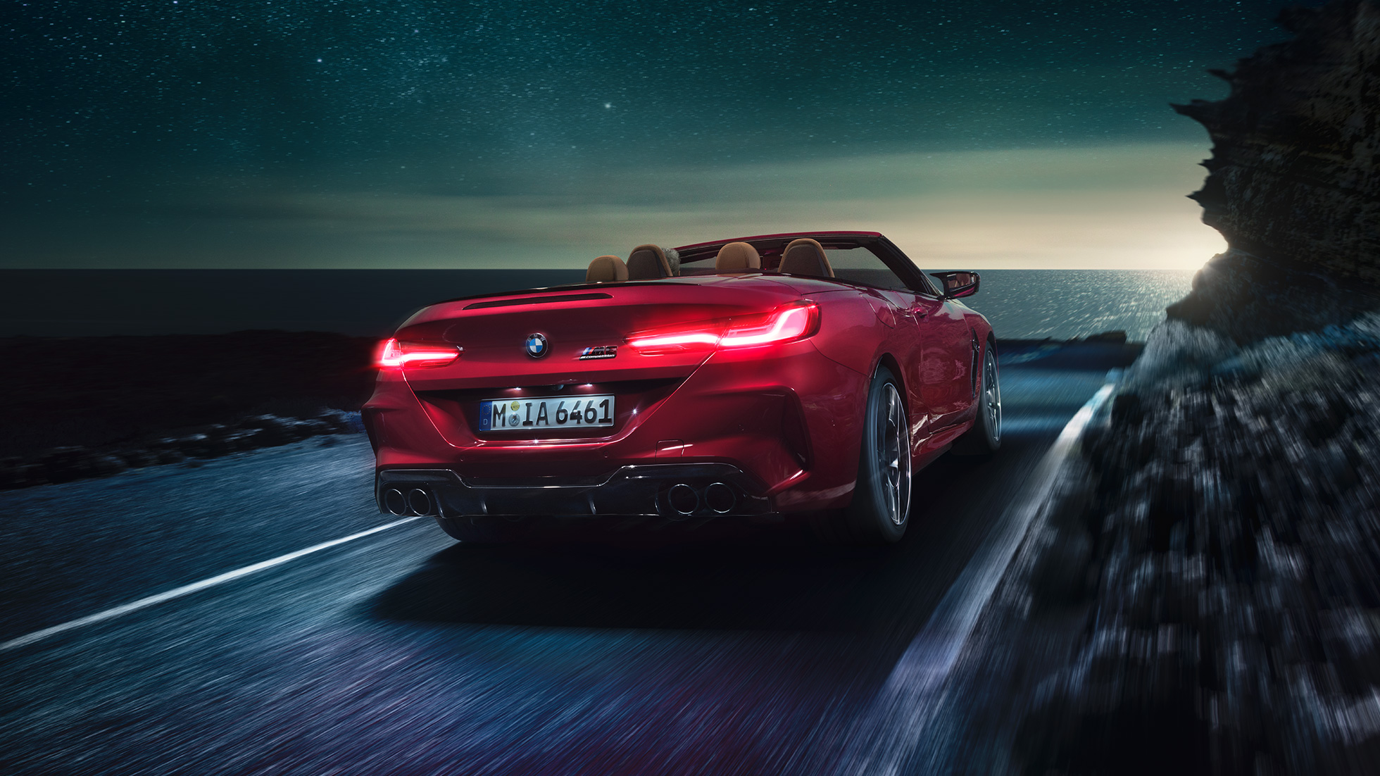 BMW M8 M Competition Cabrio, trīs ceturtdaļu skats no aizmugures. BMW M8 Competition Cabrio metāliskā Motegi Red krāsā ar nolaistu mīkstā seguma jumtu, trīs ceturtdaļu skats no aizmugures, braucošs automobilis pa piekrastes ceļu naktī.