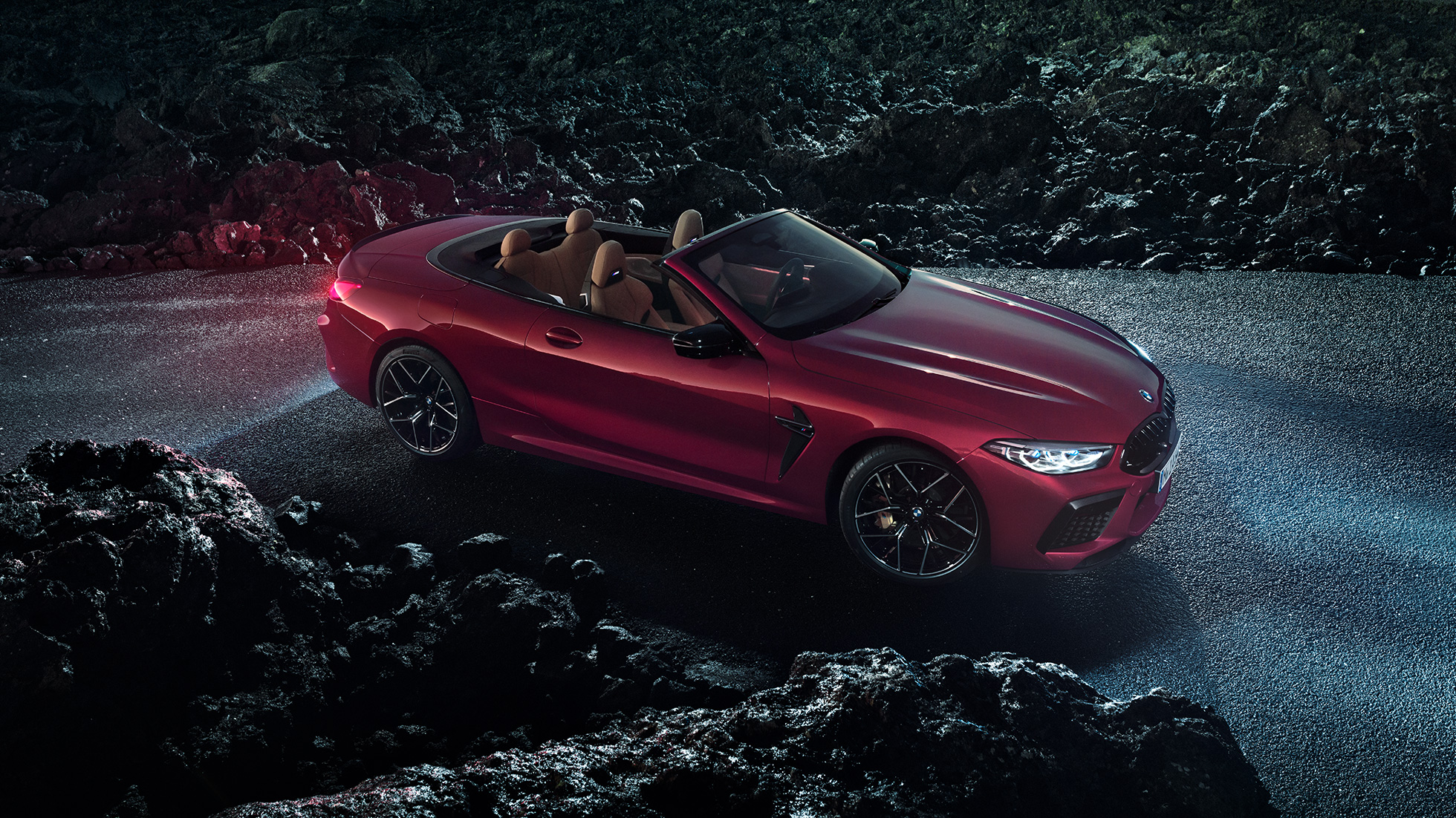 BMW M8 M Competition Cabrio, trīs ceturtdaļu skats no sāniem. BMW M8 Competition Cabrio metāliskā Motegi Red krāsā ar nolaistu mīkstā seguma jumtu, trīs ceturtdaļu skats no sāniem, stāvošs automobilis uz asfalta seguma naktī.