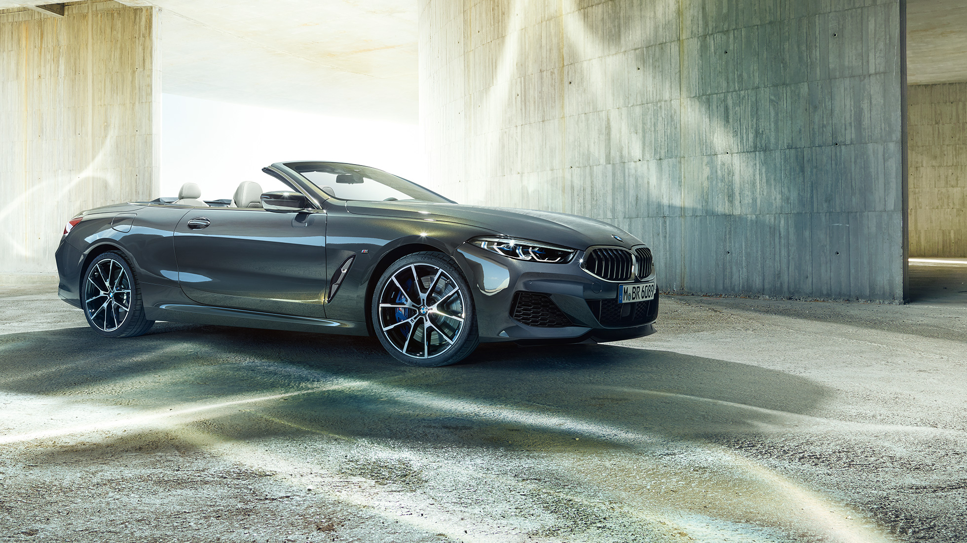 BMW M850i xDrive Cabrio, trīs ceturtdaļu skats no sāna. BMW M850i xDrive Competition Cabrio metāliskā Dravit Grey krāsā ar nolaistu mīkstā seguma jumtu, trīs ceturtdaļu skats no sāniem, stāvošs automobilis uz betona ēkas fona.