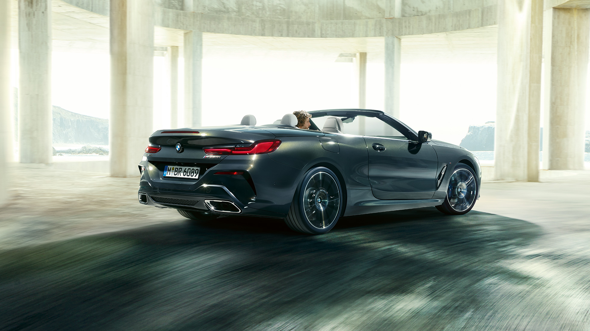 BMW M850i xDrive Cabrio, trīs ceturtdaļu skats no aizmugures. BMW M850i xDrive Competition Cabrio metāliskā Dravit Grey krāsā ar nolaistu mīkstā seguma jumtu, trīs ceturtdaļu skats no aizmugures uz automobili, kas brauc cauri betona ēkai.
